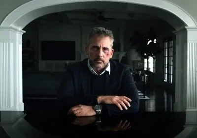 Homem de meia-idade com rosto machucado e expressão séria senta-se à mesa. Fundo de casa elegante, sombrio. Clima dramático.