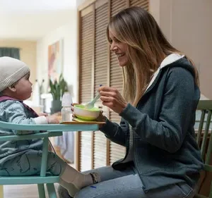 Mãe sorridente alimenta bebê em cadeira alta numa cozinha iluminada. Clima doméstico e acolhedor.