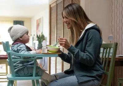 Mãe sorridente alimenta bebê em cadeira alta numa cozinha iluminada. Clima doméstico e acolhedor.
