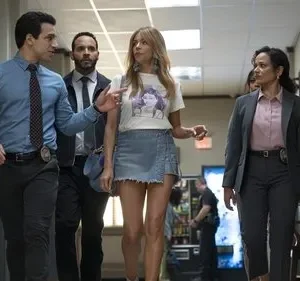 Quatro pessoas em um corredor: dois oficiais, uma mulher de camiseta e minissaia, e outra em terno. Um oficial gesticula.