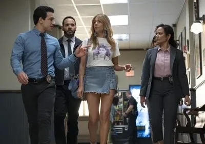 Quatro pessoas em um corredor: dois oficiais, uma mulher de camiseta e minissaia, e outra em terno. Um oficial gesticula.