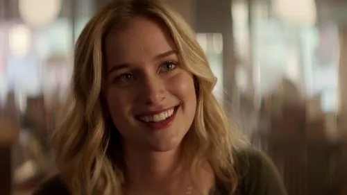 Você – T01E01:Piloto 1 Close-up de mulher loira sorridente,de olhos claros,em ambiente interno desfocado. Clima amigável e acolhedor.