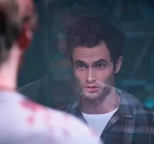 Homem de cabelo escuro e camisa xadrez olha fixamente pessoa de costas com sangue na roupa. Um vidro os separa, gerando tensão.