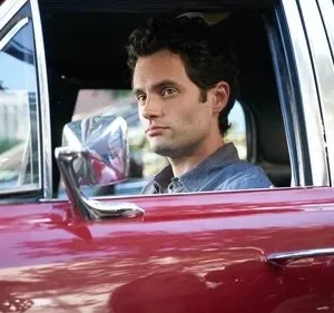 Um homem de cabelo escuro e olhar intenso está sentado ao volante de um carro vermelho-escuro, olhando para o lado.
