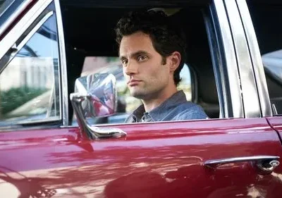 Um homem de cabelo escuro e olhar intenso está sentado ao volante de um carro vermelho-escuro, olhando para o lado.