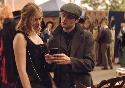 Mulher de espartilho preto e homem de boné olham um celular em um mercado movimentado com pessoas em trajes de época.