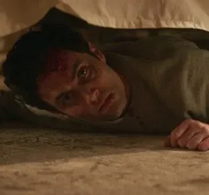 Homem ferido com sangue na testa e olhar fixo, deitado no chão sob uma cama. Um copo de bebida perto. Ambiente perturbador.