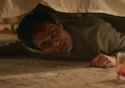 Homem ferido com sangue na testa e olhar fixo, deitado no chão sob uma cama. Um copo de bebida perto. Ambiente perturbador.