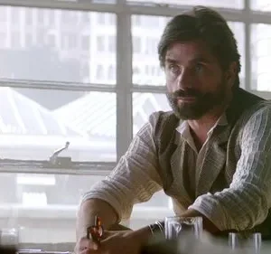 Homem barbudo de colete sentado em poltrona de couro, olhando pensativo. Fundo com janelas de escritório e cidade.