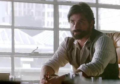 Homem barbudo de colete sentado em poltrona de couro, olhando pensativo. Fundo com janelas de escritório e cidade.