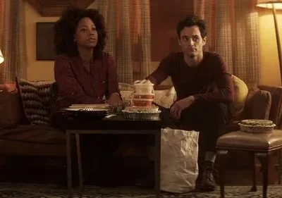 Mulher e homem sentados em sala com pouca luz. Uma mesa baixa com comida para viagem está entre eles. Ambos têm expressões pensativas.