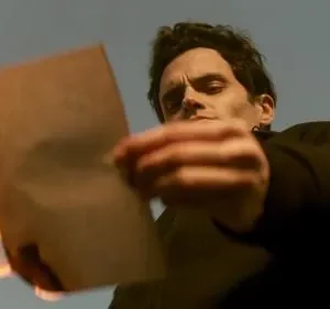 Um homem de cabelo escuro olha fixamente um papel em chamas, contra um céu azul. Ação intensa e dramática.