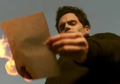 Um homem de cabelo escuro olha fixamente um papel em chamas, contra um céu azul. Ação intensa e dramática.