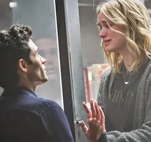 Homem observa mulher loira chorando através de um vidro. Fundo de estantes de livros. Clima de drama e confinamento.