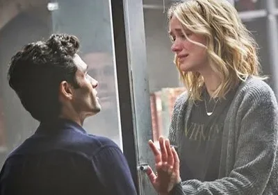 Homem observa mulher loira chorando através de um vidro. Fundo de estantes de livros. Clima de drama e confinamento.