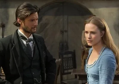Um homem barbudo de terno escuro e uma mulher de vestido azul em cenário rústico. Ambos parecem sérios e pensativos.