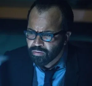 Um homem de barba e óculos olha intensamente para uma tela azulada. O ambiente escuro sugere foco e tensão.