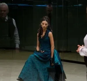 Mulher de vestido azul sentada, olhando para baixo. À direita, outra mulher de costas. Homem sério reflete-se ao fundo. Ambiente espelhado e sombrio.