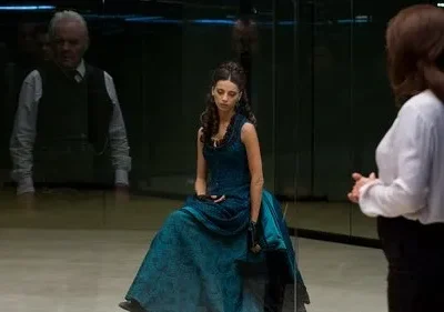 Mulher de vestido azul sentada, olhando para baixo. À direita, outra mulher de costas. Homem sério reflete-se ao fundo. Ambiente espelhado e sombrio.