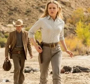 Mulher loira e homem em trajes western, com coldres, caminham por um deserto árido. Corpos jazem ao redor. Clima tenso de ação.