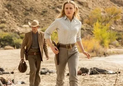 Mulher loira e homem em trajes western, com coldres, caminham por um deserto árido. Corpos jazem ao redor. Clima tenso de ação.