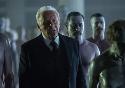 Um homem idoso de terno observa figuras masculinas sem camisa, de pele cerosa, em ambiente escuro e misterioso.