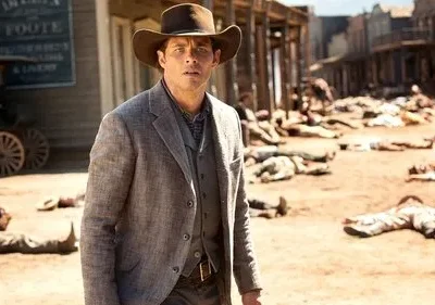 Homem de chapéu cowboy e terno olha chocado uma rua empoeirada de Velho Oeste, com corpos espalhados.