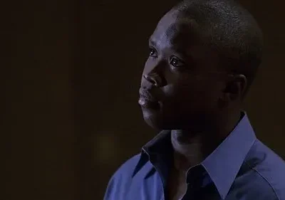 Jovem negro de camisa azul, olhando para cima com expressão pensativa. Fundo escuro e difuso cria um clima sombrio.