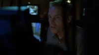 Homem de cabelo claro,olhar atento,em sala escura. Luz azul/verde ilumina seu rosto. Monitores no fundo. Clima de suspense.