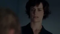 Mulher de cabelo escuro com expressão preocupada,olhando para a esquerda em um ambiente escuro e tenso.