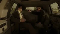 Dentro de um carro,um homem de terno tem uma pistola apontada para a cabeça por outro homem de óculos escuros. Cena tensa.
