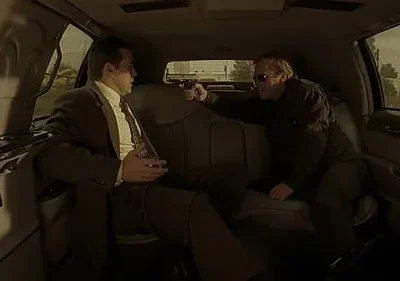 Dentro de um carro, um homem de terno tem uma pistola apontada para a cabeça por outro homem de óculos escuros. Cena tensa.