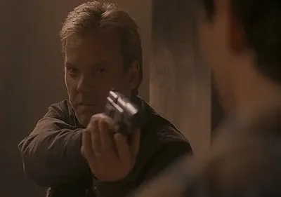 Homem loiro de expressão séria aponta pistola para figura desfocada à direita. Ambiente escuro e esfumaçado. Cena de ação tensa.