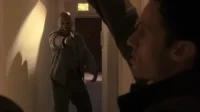 Num corredor escuro,um homem careca e irritado aponta uma pistola diretamente. Outro homem,em primeiro plano,reage com surpresa e medo.