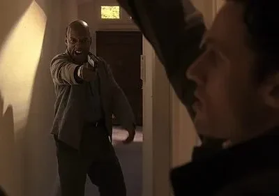 Num corredor escuro, um homem careca e irritado aponta uma pistola diretamente. Outro homem, em primeiro plano, reage com surpresa e medo.