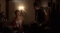 Mulher assustada olha para homem em sala escura. Garrafas e pouca luz criam clima tenso.