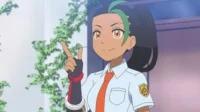 Anime: jovem morena sorrindo, cabelo preto c/ mechas verdes, camisa escolar, gravata laranja, luvas. Aponta p/ cima em fundo claro com plantas.