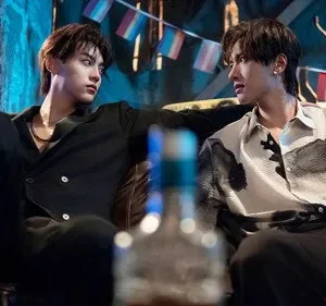 Dois jovens sentados num bar escuro. Um de preto olha fixamente para o outro, que veste camisa estampada e parece tenso. Bandeiras LGBTQ+ ao fundo.