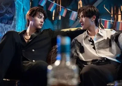 Dois jovens sentados num bar escuro. Um de preto olha fixamente para o outro, que veste camisa estampada e parece tenso. Bandeiras LGBTQ+ ao fundo.