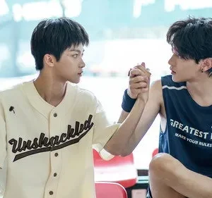 Dois rapazes jovens se encaram com as mãos unidas em uma quadra. Um de camisa beisebol creme, outro de regata azul. Clima íntimo.