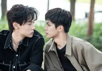 Dois jovens olham-se intensamente ao ar livre. Um veste camisa preta, o outro casaco cáqui. Expressões sérias em ambiente natural.