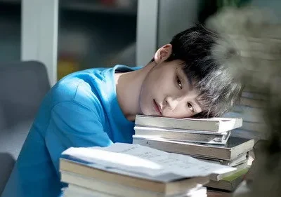 Garoto asiático de camiseta azul, deitado sobre livros e papéis, com olhar cansado sob a luz do sol. Ambiente de estudo.