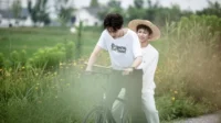 Dois jovens em uma bicicleta em um campo verde. Um sorri, de chapéu de palha, o outro pedala. Cena alegre e de verão.