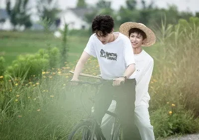 Dois jovens em uma bicicleta em um campo verde. Um sorri, de chapéu de palha, o outro pedala. Cena alegre e de verão.
