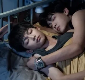 Dois jovens asiáticos dormem abraçados em uma cama. A cena é íntima e serena, com iluminação fraca.