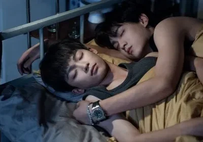 Dois jovens asiáticos dormem abraçados em uma cama. A cena é íntima e serena, com iluminação fraca.