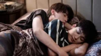 Dois jovens dormindo abraçados em uma cama. Um de camisa preta, outro listrada. Lençóis com estampa de onça. Cena íntima e serena.