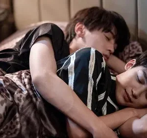 Dois jovens dormindo abraçados em uma cama. Um de camisa preta, outro listrada. Lençóis com estampa de onça. Cena íntima e serena.