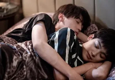 Dois jovens dormindo abraçados em uma cama. Um de camisa preta, outro listrada. Lençóis com estampa de onça. Cena íntima e serena.