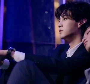 Dois homens jovens em ternos sob luz azul. Um olha para cima, o outro dorme em seu ombro. Clima íntimo e melancólico.